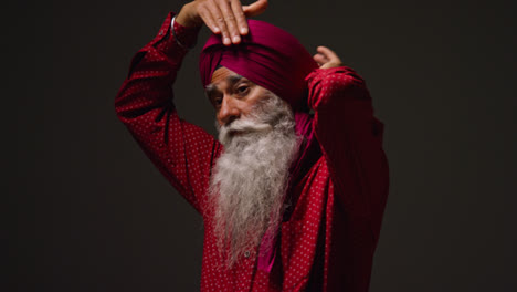 Low-Key-Studio-Lighting-Shot-Of-Senior-Sikh-Man-With-Beard-Tying-Fabric-For-Turban-And-Using-Salai-Needle-Against-Dark-Background